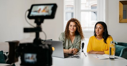 Two people sit on either side of a microphone, recording a video. One sit with a laptop in front of them while the other has a notebook and water bottle.