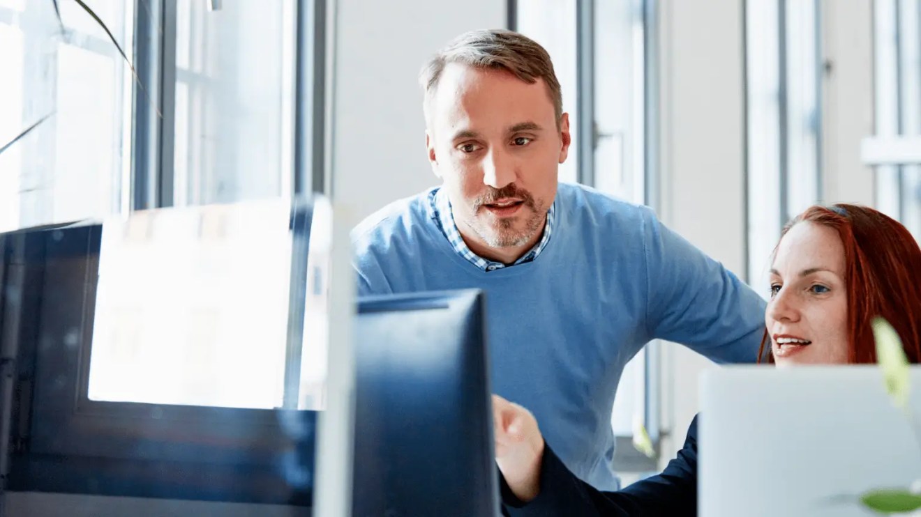 Two coworkers reviewing content and discussing a digital solution dashboard on a computer monitor.