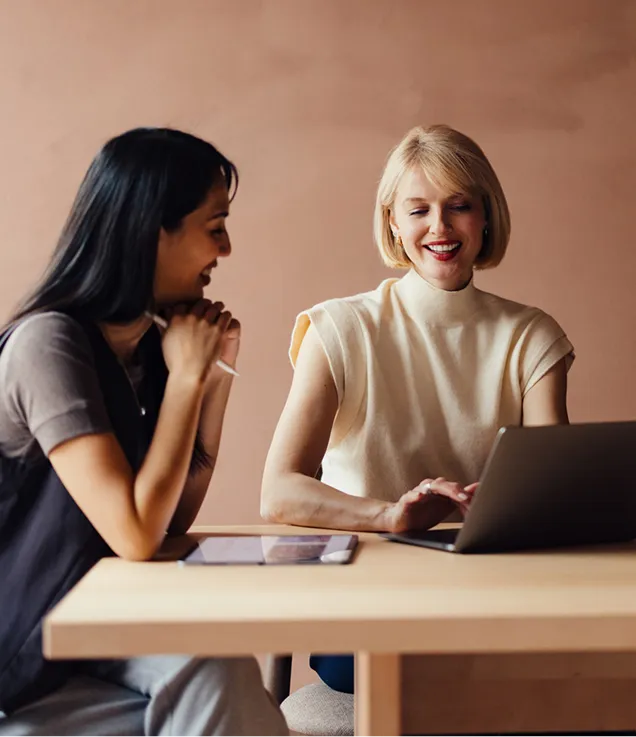 Two colleagues smiling while reviewing content together on a laptop at a meeting table, with a tablet placed beside them.