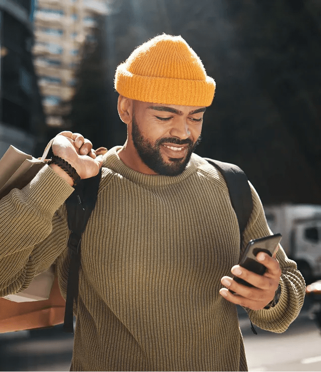 Smiling man in a yellow beanie checks his phone while carrying shopping bags outdoors.