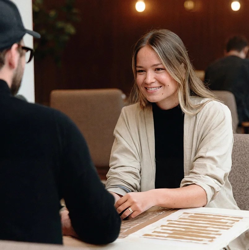 Two people seated across a table, smiling during a conversation, with printed documents laid out in front of them.