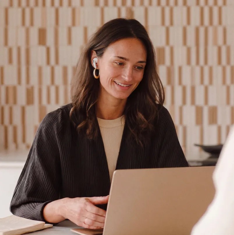 Person wearing wireless earbuds smiling while participating in a video call on a laptop at a desk.
