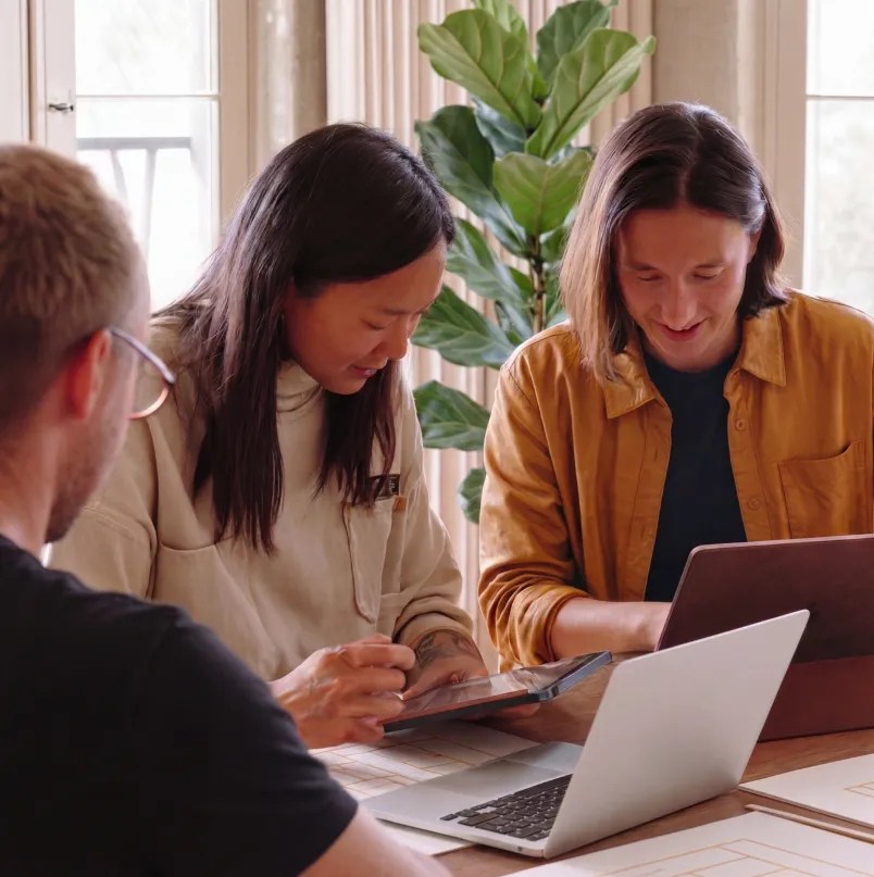 Three colleagues collaborating around a table, using a laptop and tablet to review work as part of WordPress VIP solutions for cross-functional teams.