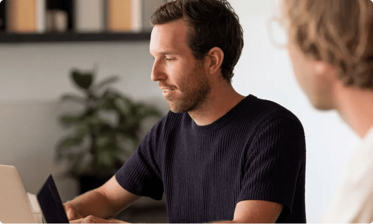 Two people sit at a table working on laptops, with shelves and a plant in the background—collaborating seamlessly as they explore insights provided by a content analytics AI assistant.