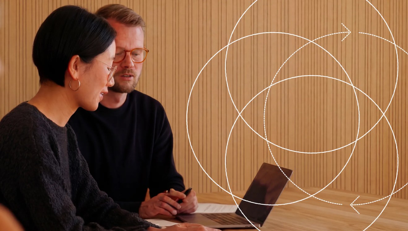 Two people sitting at a table, looking at a laptop, with abstract white lines over a wooden wall—collaborating on a digital project powered by headless WordPress CMS.