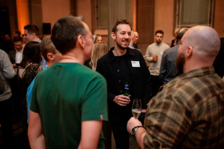 A group of people are socializing indoors at an Innovation Exchange event, some holding drinks and smiling while talking.