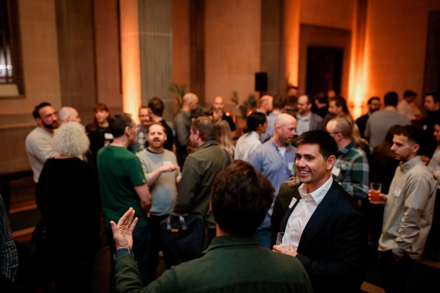A group of people socialize at an indoor Innovation Exchange event; one man in a suit holds a drink and smiles.