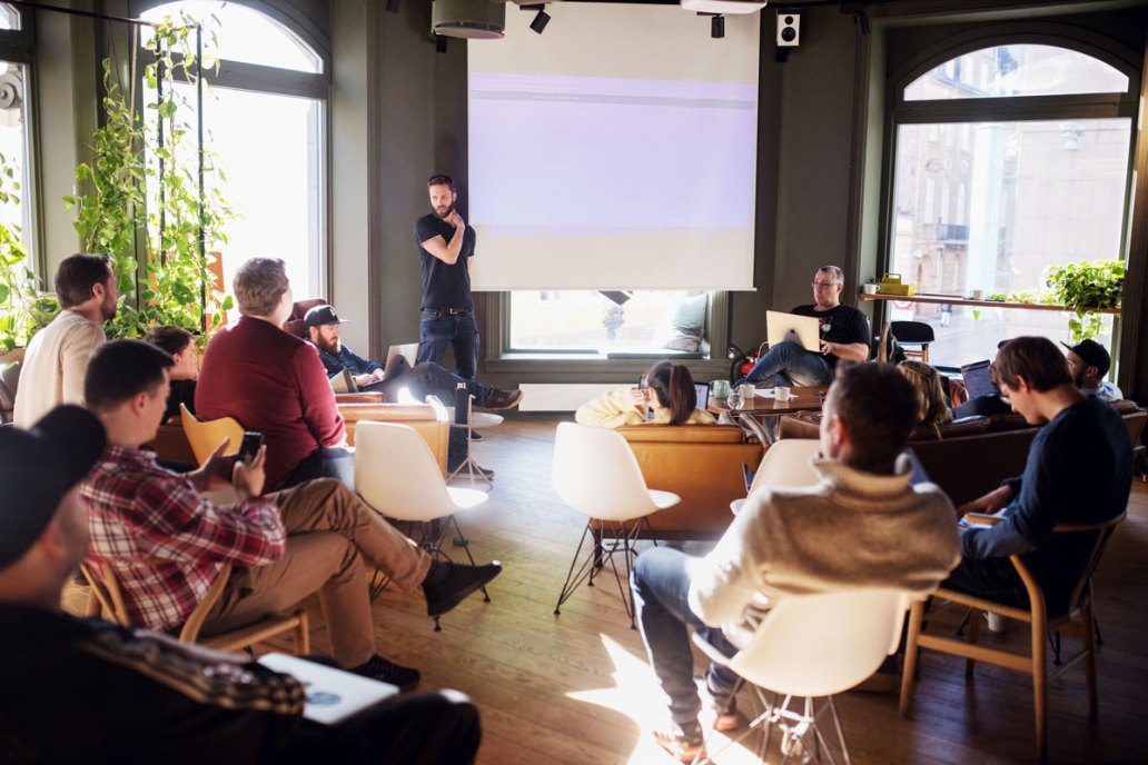 A team of people collaborating in a sunlit room, a person stands next to a blank projector screen while other sit working on various devices