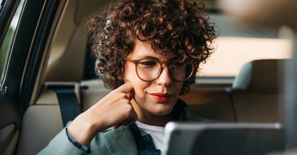 A person with a smirking at a tablet screen with glasses on while in the back seat of a car