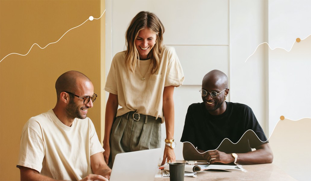 Three colleagues smiling and collaborating around a laptop at a desk.